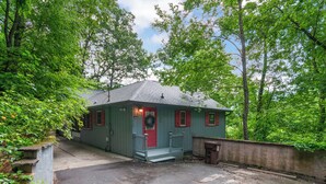 Eine kleine, eingeschossige Holzhütte mit roter Tür und einer Veranda, umgeben von dichtem Grün.