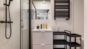 Un baño moderno con cabina de ducha de vidrio, un lavabo blanco con artículos de aseo, un perchero negro y un estante negro.