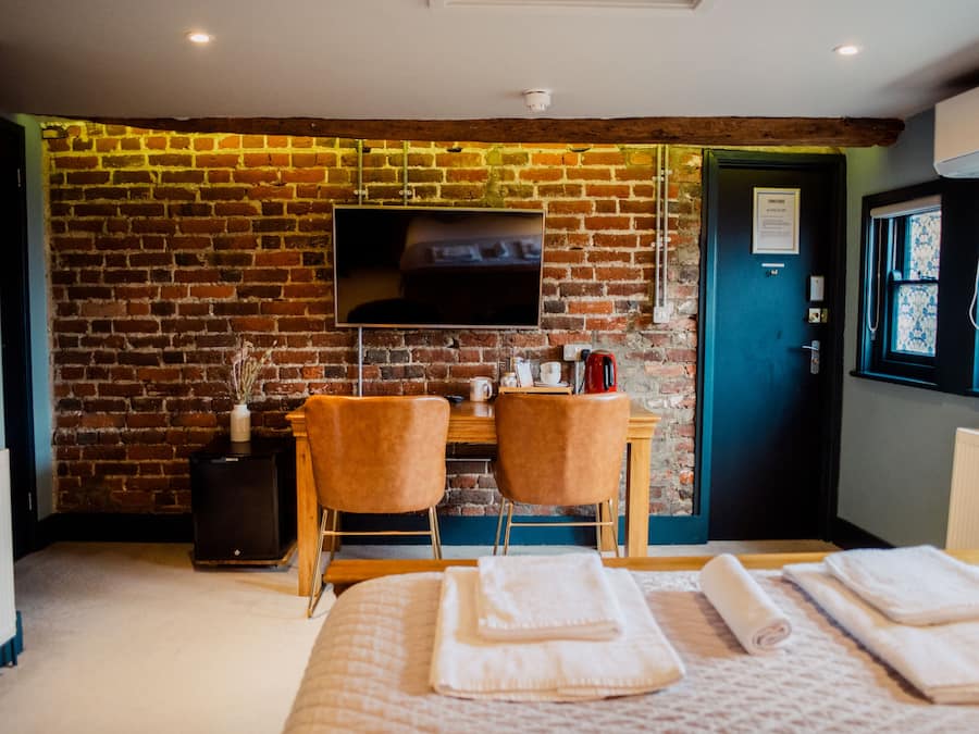 Une pièce avec un mur en briques, un téléviseur à écran plat, deux chaises, un bureau, un lit avec des serviettes et une porte avec une fenêtre.