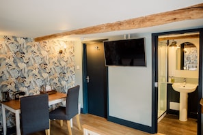 A room with a wooden table, two chairs, a TV mounted on the wall, a door, and a bathroom with a sink and mirror. - Corner House (Canterbury)