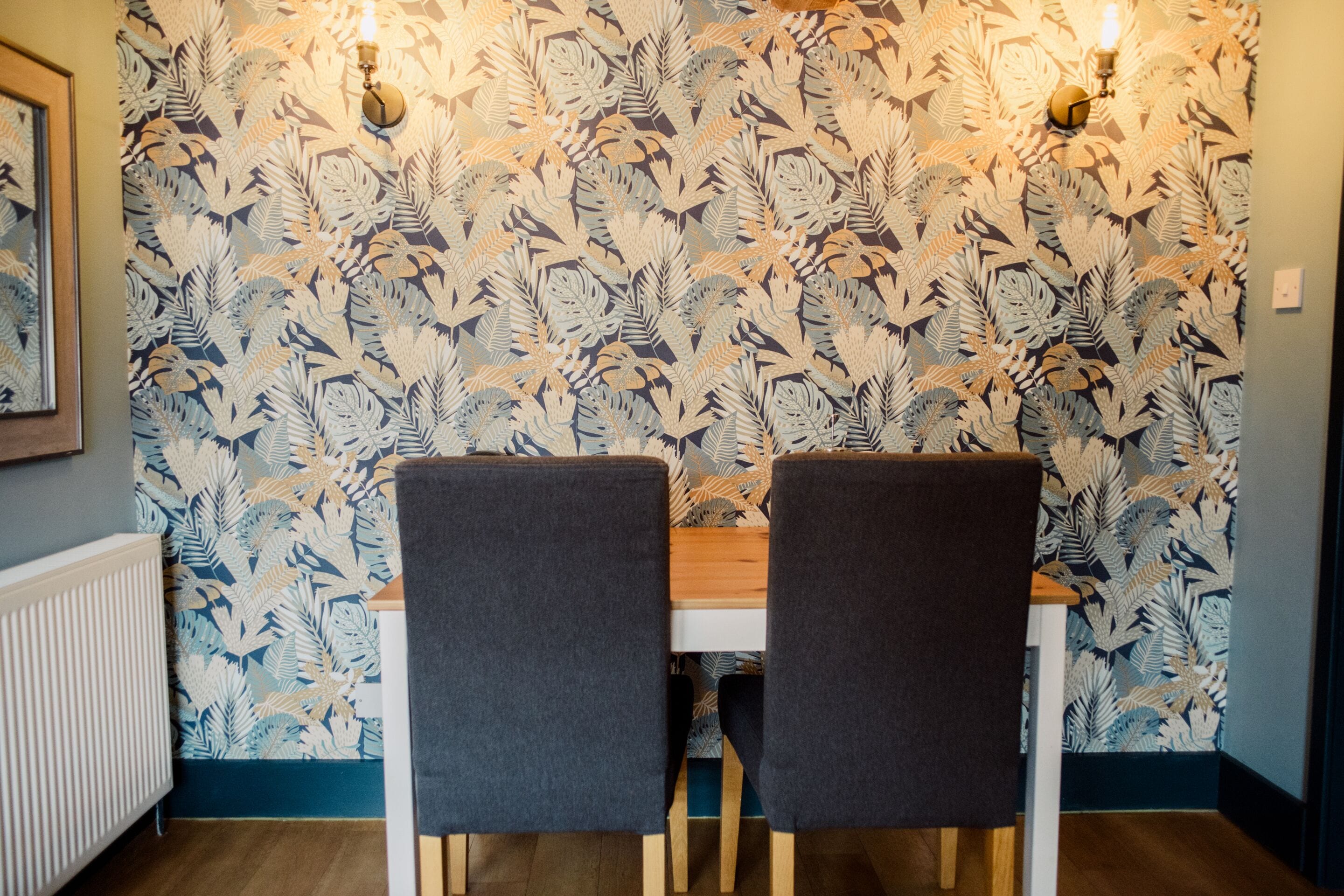 Un comedor con papel tapiz estampado, una mesa, dos sillas y luces fijadas a la pared.