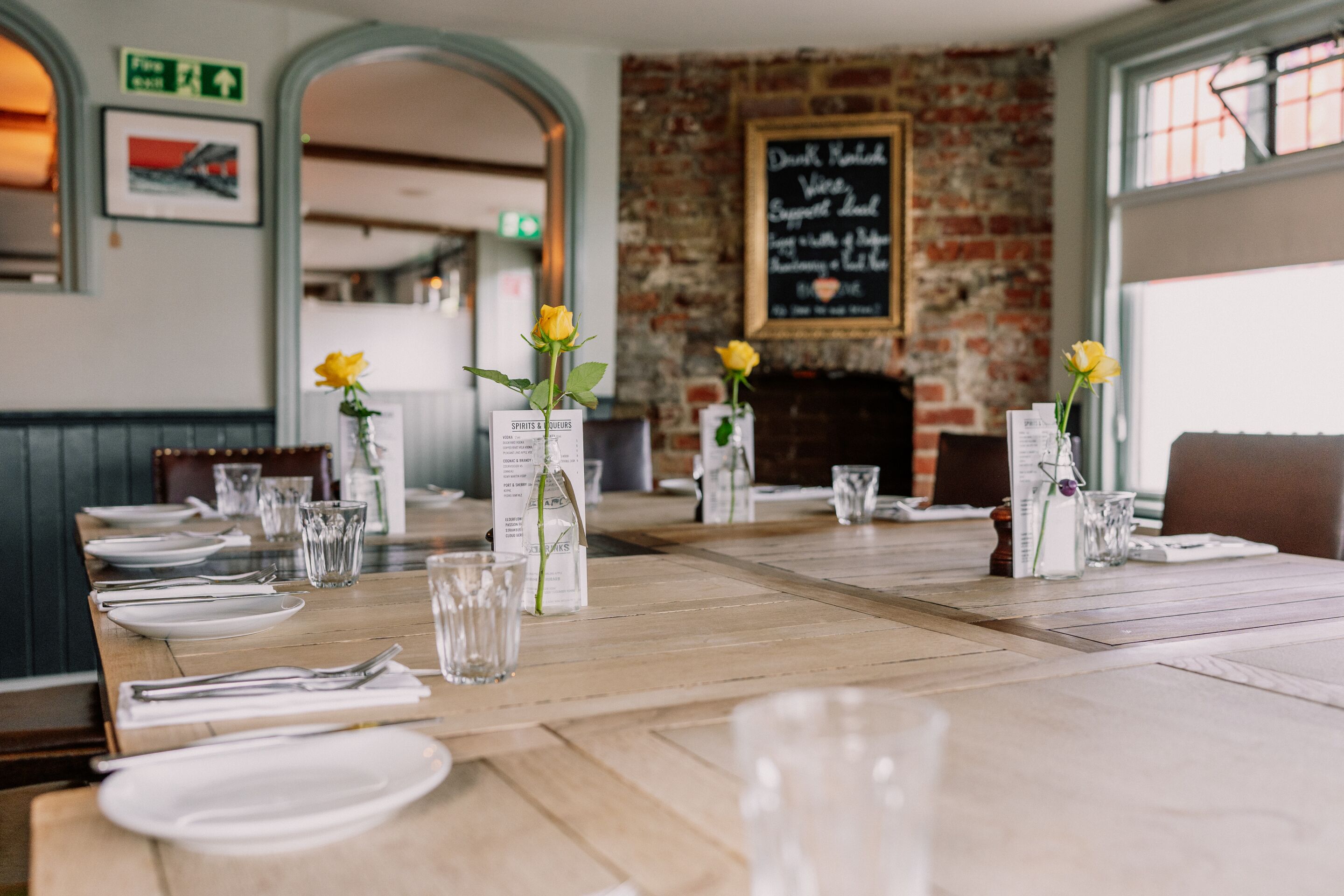 A restaurant with wooden tables, yellow roses in vases, and a brick wall with a chalkboard menu.