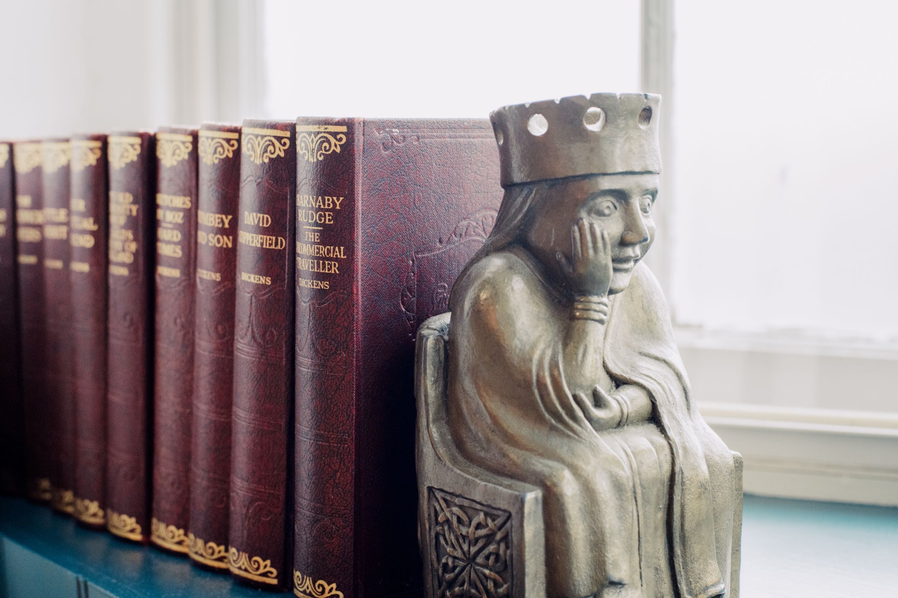 Una hilera de libros con letras doradas en el lomo, una estatua de una figura sentada con corona y una ventana al fondo.