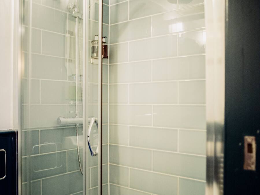 Une douche fermée par des parois de verre, avec des carreaux blancs, une douchette à main et une porte en verre.