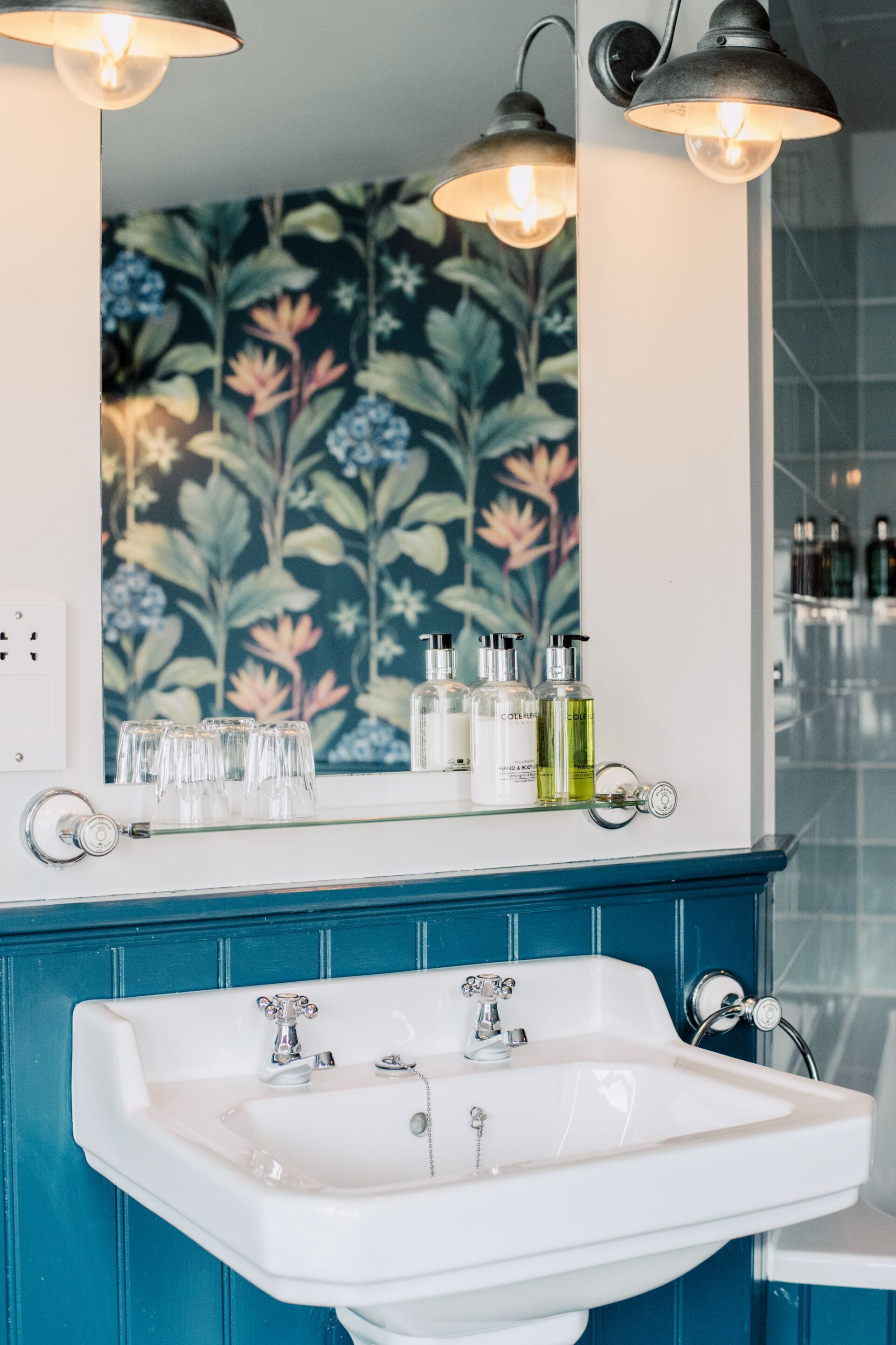 Un baño con lavamanos blanco, muebles de color azul, papel tapiz floral y dos luces fijadas a la pared.