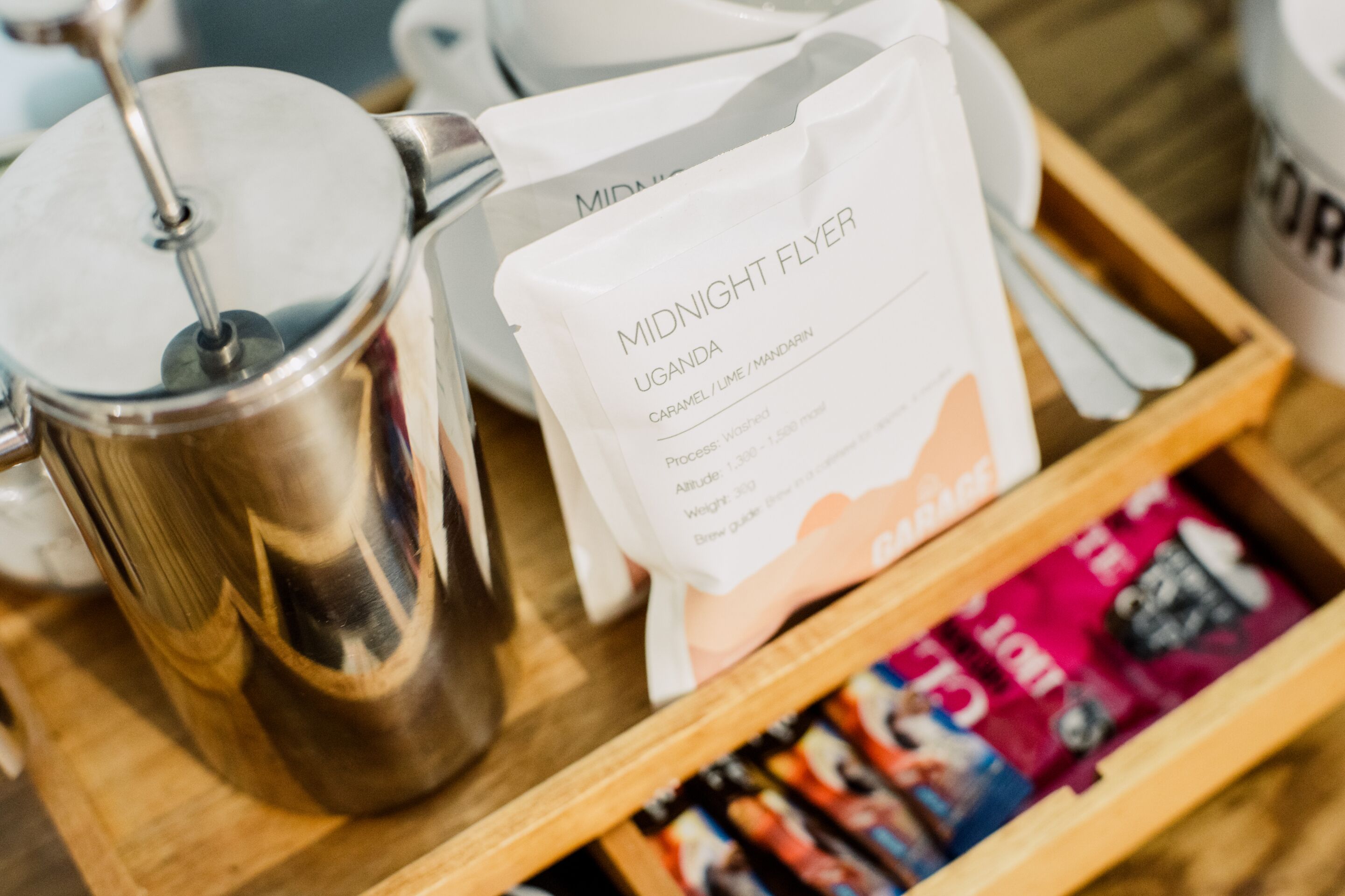 A coffee maker, a bag labeled 'Midnight Flyer Uganda Caramel/Lime/Mandarin,' and a wooden tray with coffee packets.