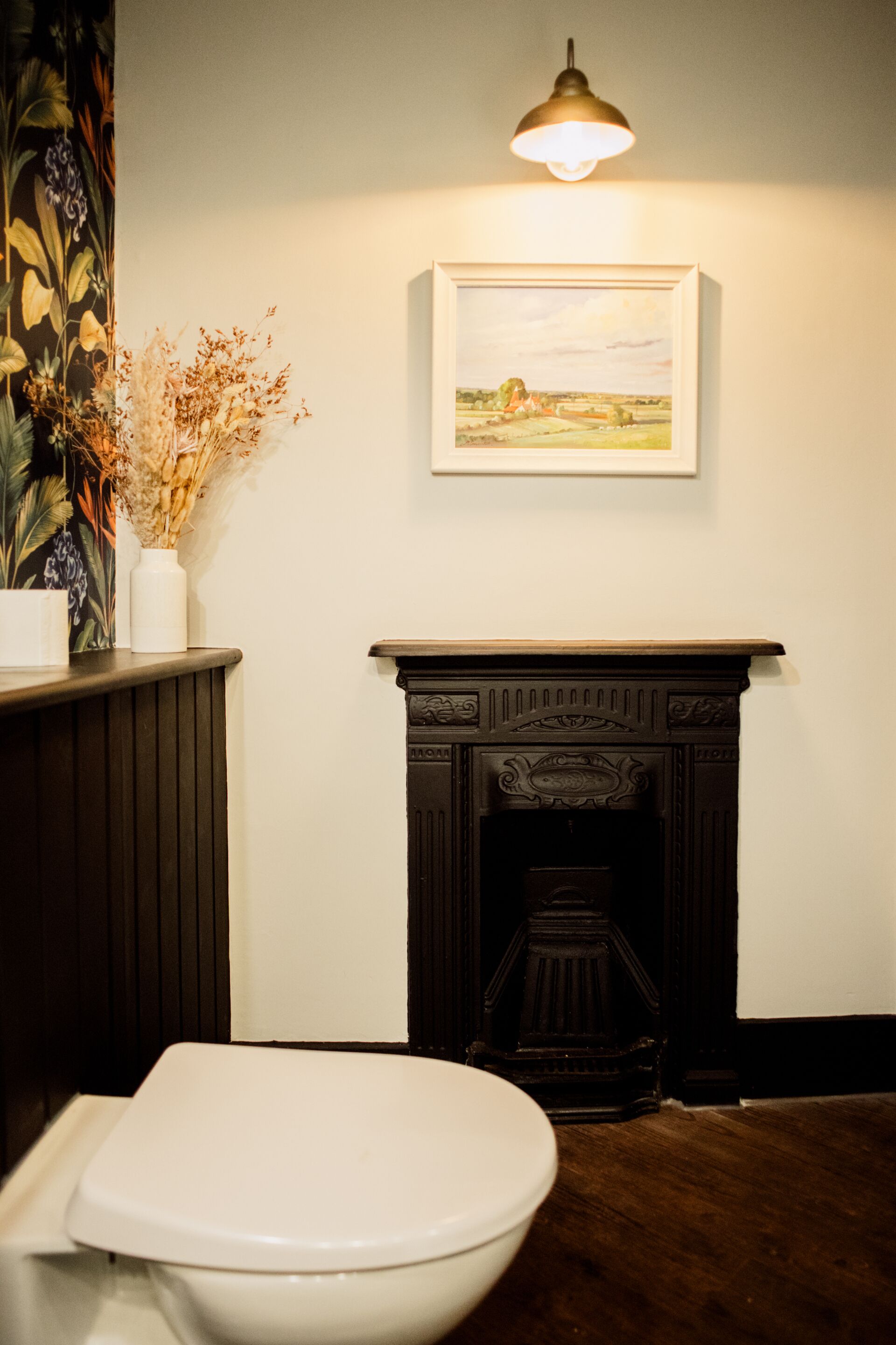 A bathroom with a toilet, a wall-mounted light fixture, a framed painting, and a fireplace.