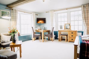 Une pièce comprenant un bureau en bois, deux chaises, un téléviseur, une petite table avec un vase de fleurs, un canapé et un lit recouvert d’une couverture à carreaux.