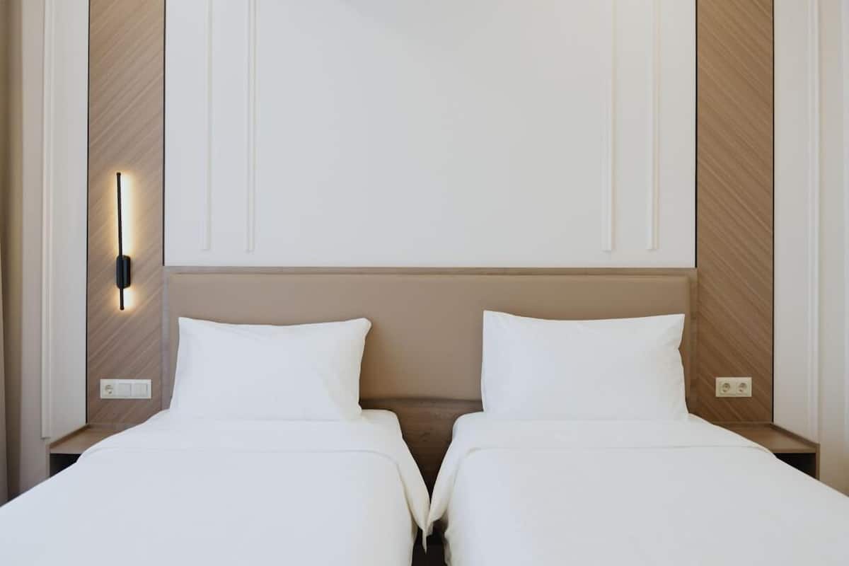 A hotel room with two beds, a nightstand with a lamp, and a wall-mounted light fixture.