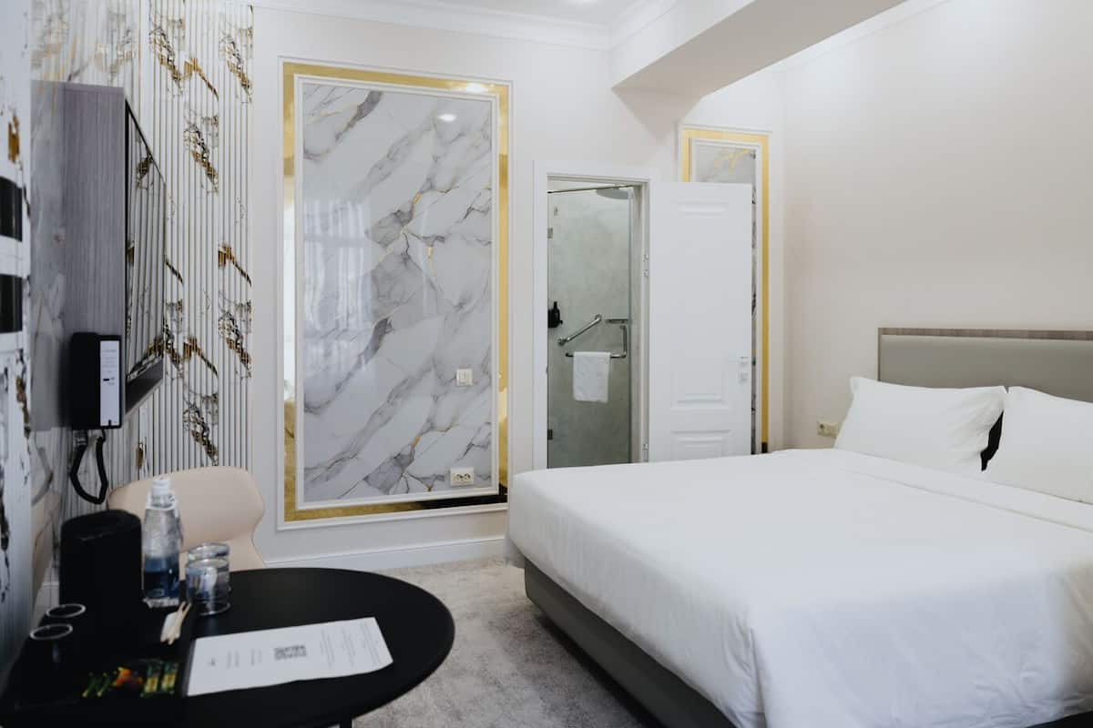 A hotel room with a large bed, a desk with a chair, a minibar, and a bathroom with a glass door.