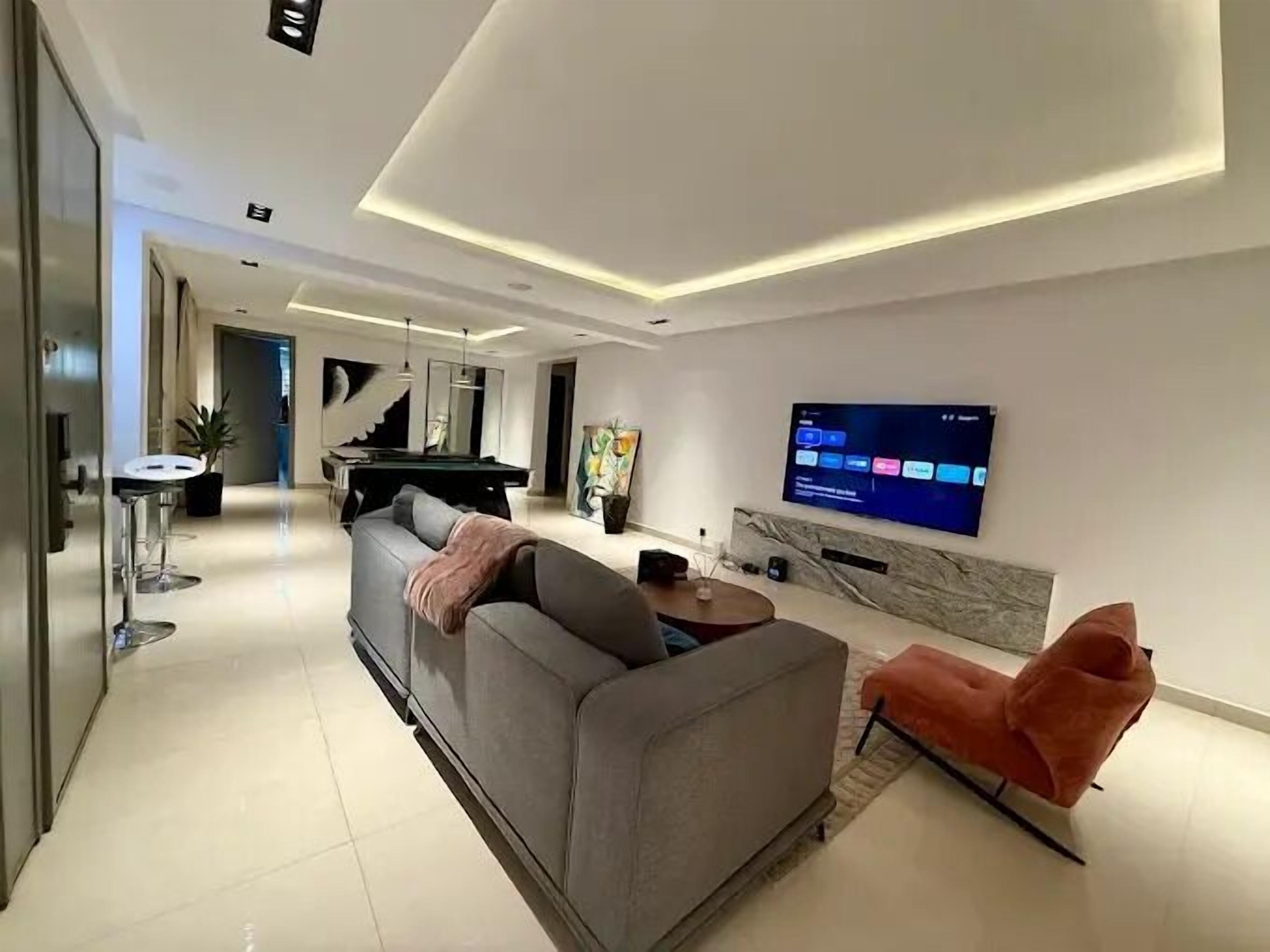 A modern living room with a grey sofa, a pool table, and a flat-screen TV mounted on the wall.