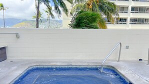 A rectangular swimming pool with a metal ladder, surrounded by a white wall and palm trees. - Aloha Living at Waikiki Lanais | Pool + Gym (Honolulu)
