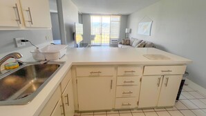 A modern kitchen with white cabinets, a large island, and a view to the outside. - Aloha Living at Waikiki Lanais | Pool + Gym (Honolulu)