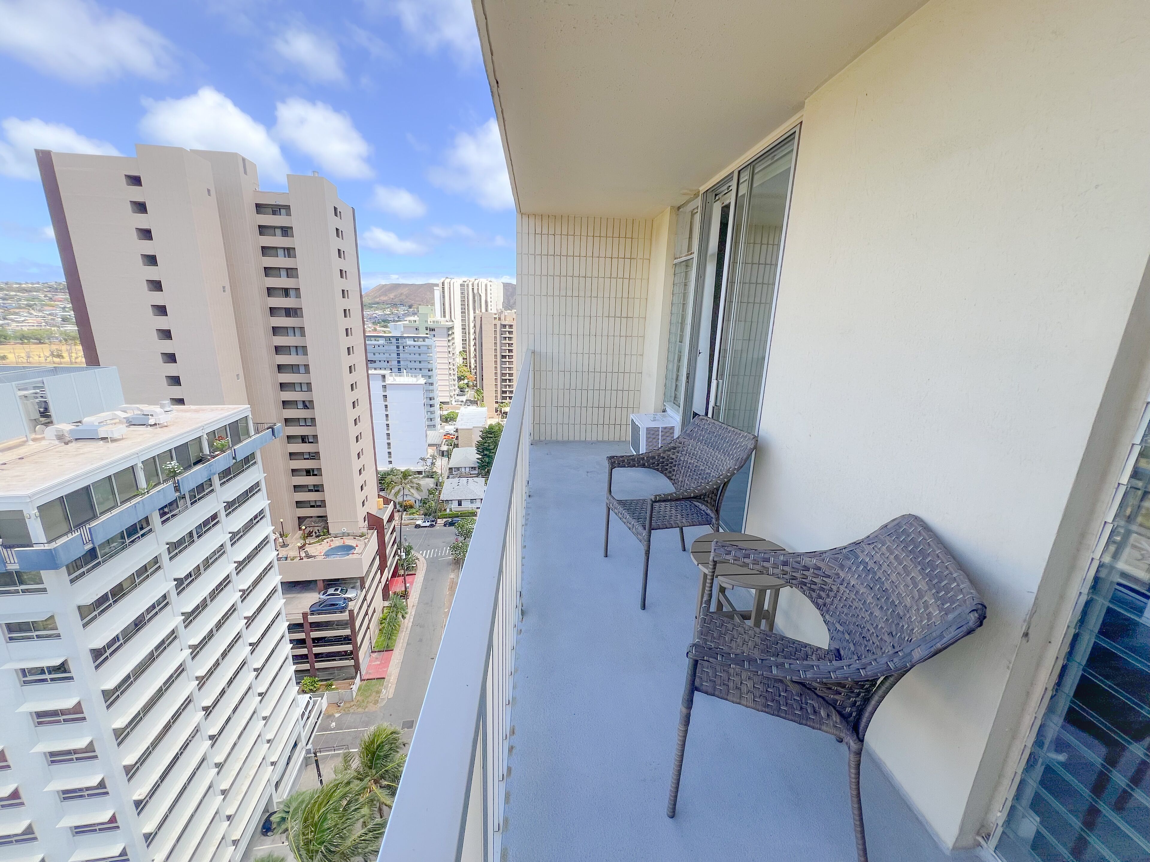 A balcony with two wicker chairs and a small table, overlooking a cityscape with tall buildings.