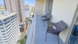 A balcony with two wicker chairs and a small table, overlooking a cityscape with tall buildings. - Aloha Living at Waikiki Lanais | Pool + Gym (Honolulu)