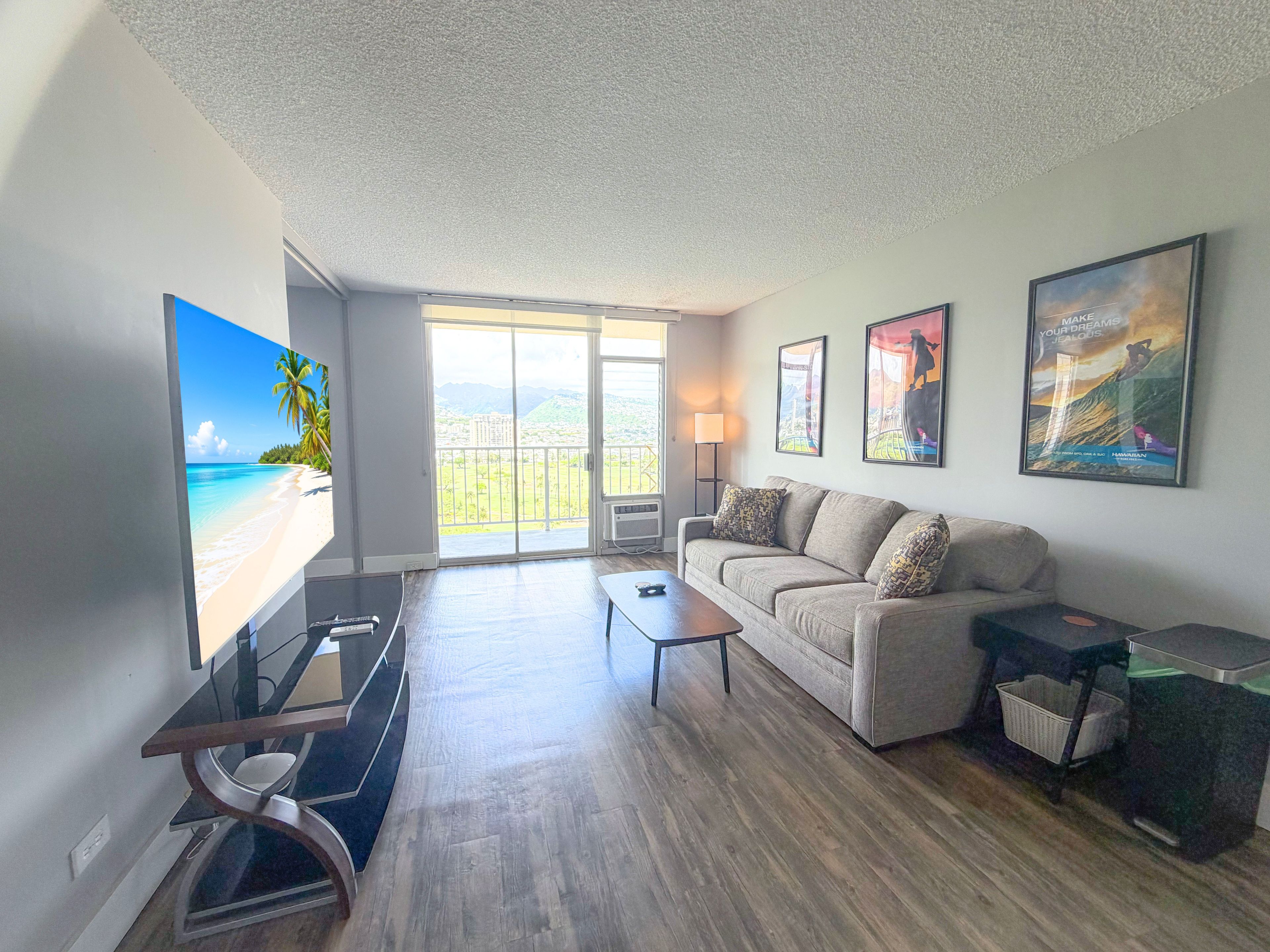 A modern living room with a large flat-screen TV, a sofa, a coffee table, and a balcony with a view of a beach and mountains.