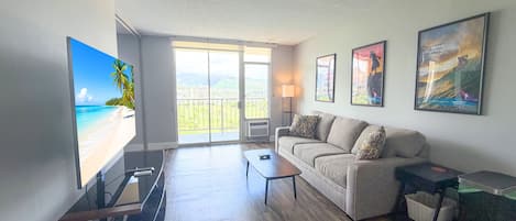 A modern living room with a large flat-screen TV, a sofa, a coffee table, and a balcony with a view of a beach and mountains.