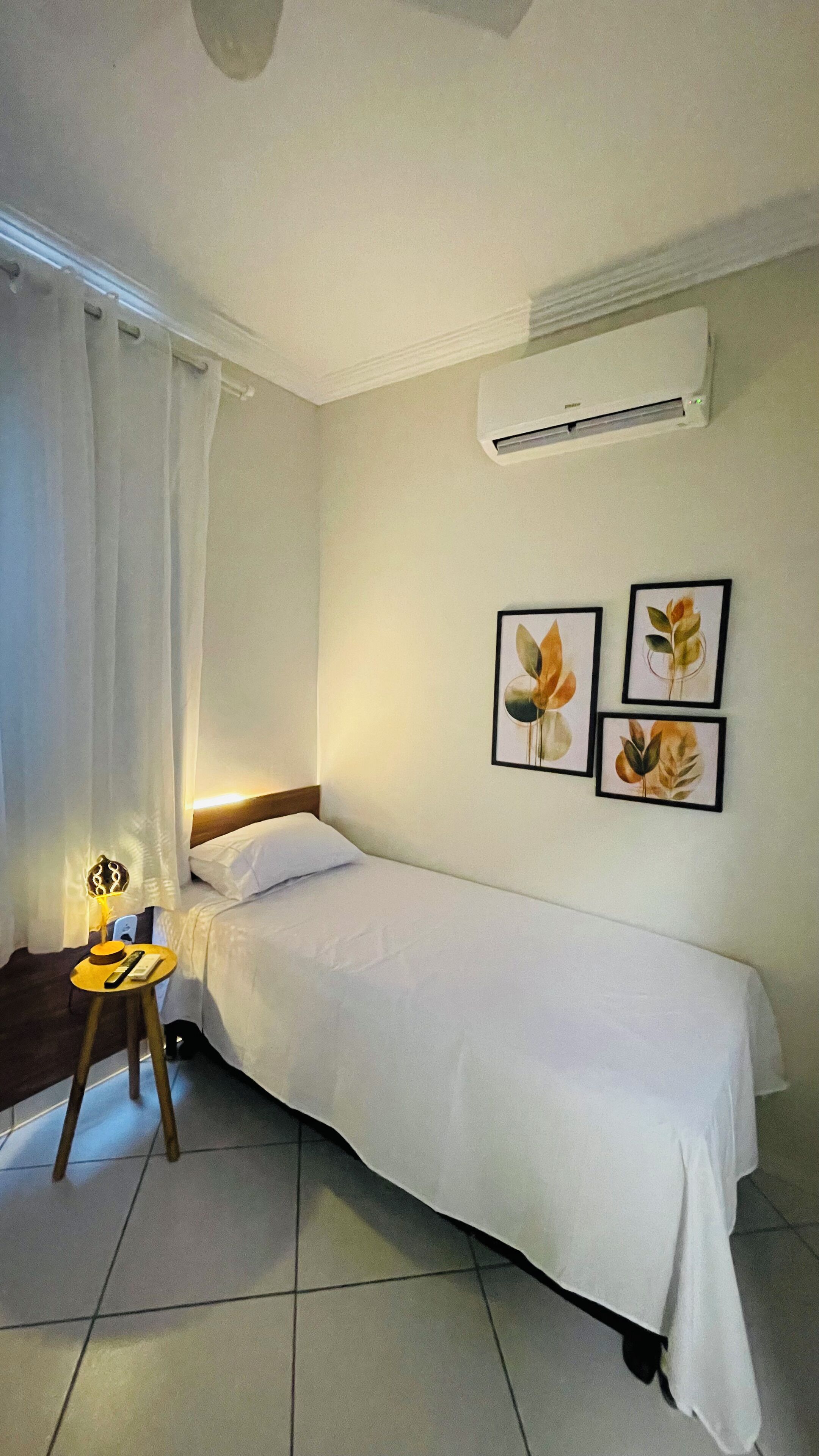 A single bed with a white bedspread, a bedside table with a lamp, and three framed leaf paintings on the wall.