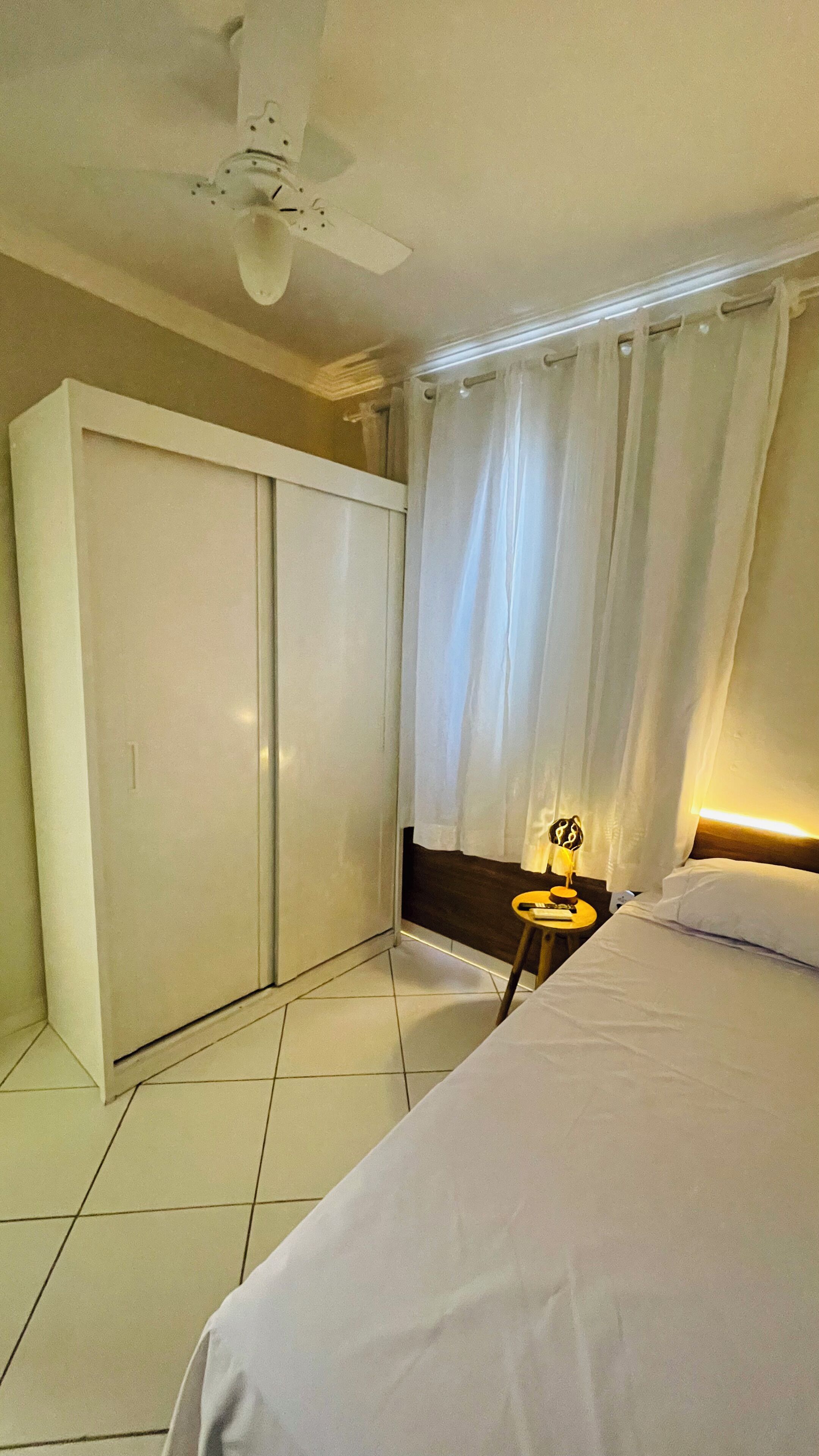 Une chambre à coucher comprenant un lit, une table de chevet, un ventilateur de plafond, une armoire et une fenêtre avec des rideaux.