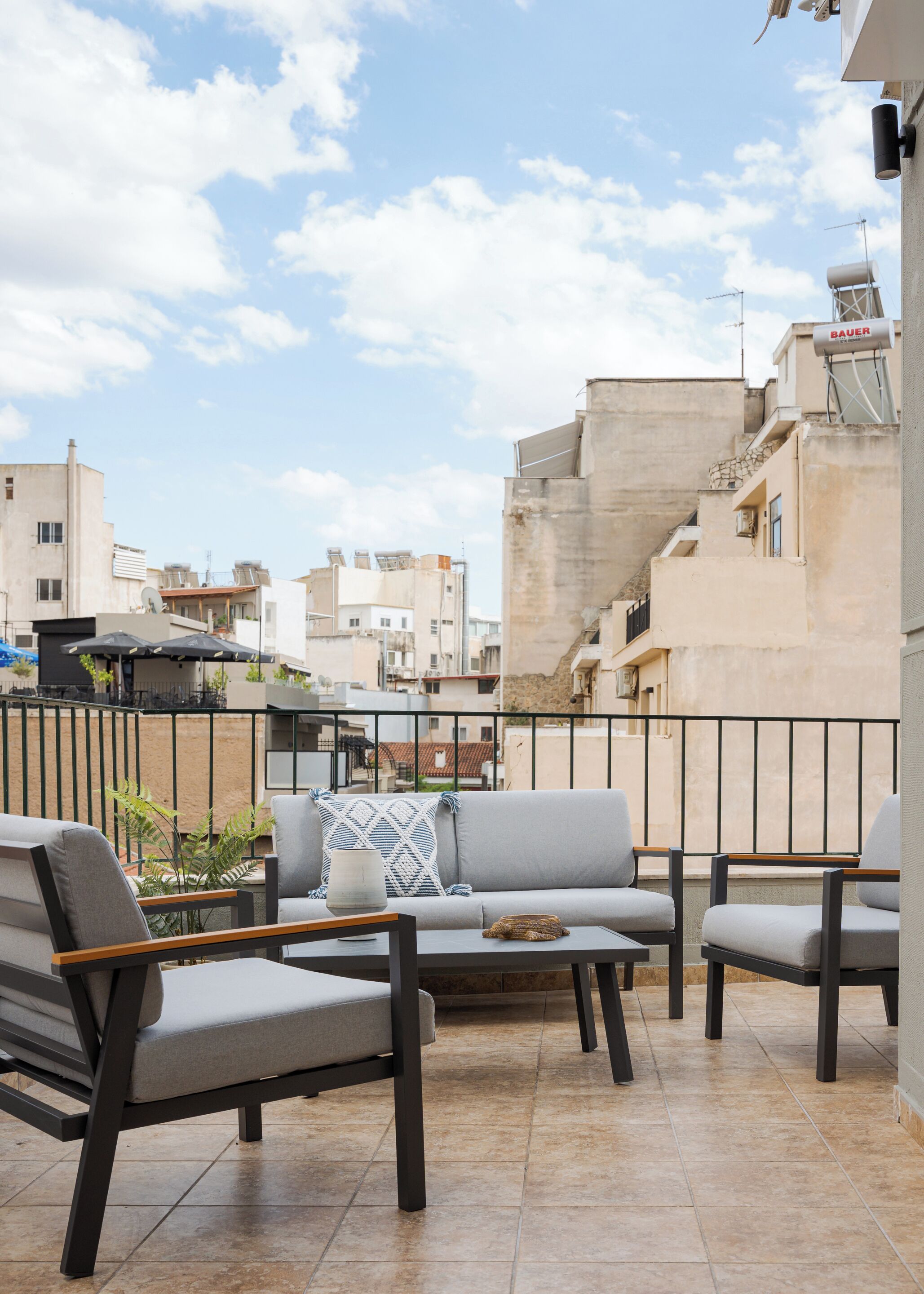 A terrace with a grey sofa, two armchairs, and a small table. The terrace has a tiled floor and a metal railing. In the background, there are buildings with balconies and a clear sky.