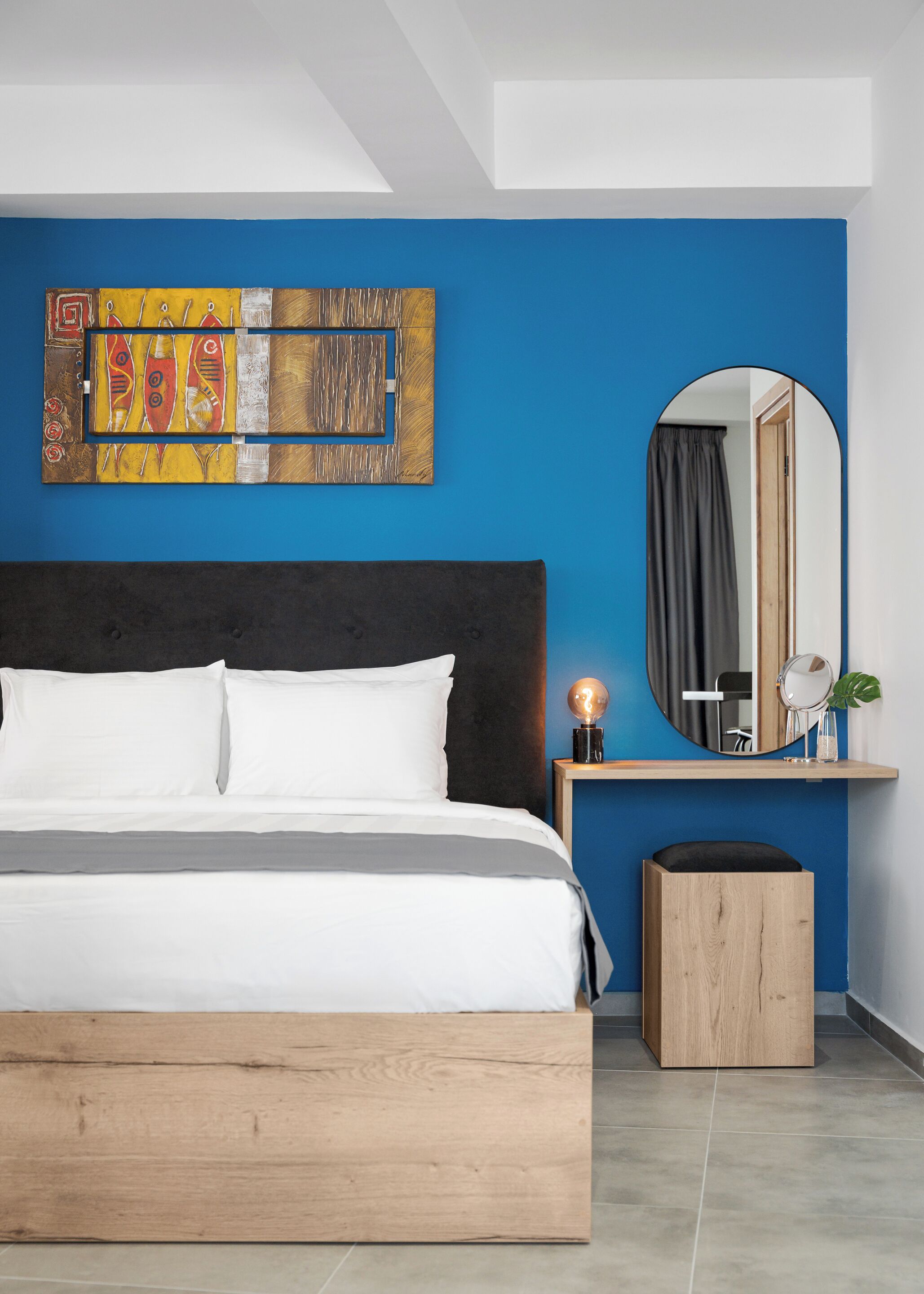 Un dormitorio moderno con una cama grande, cabecera de madera, pared de acento azul, espejo montado en la pared y una mesita de noche con lámpara.