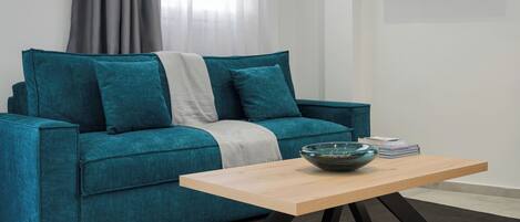 A teal sofa with a white throw blanket, a wooden coffee table with a bowl, and a TV mounted on the wall.