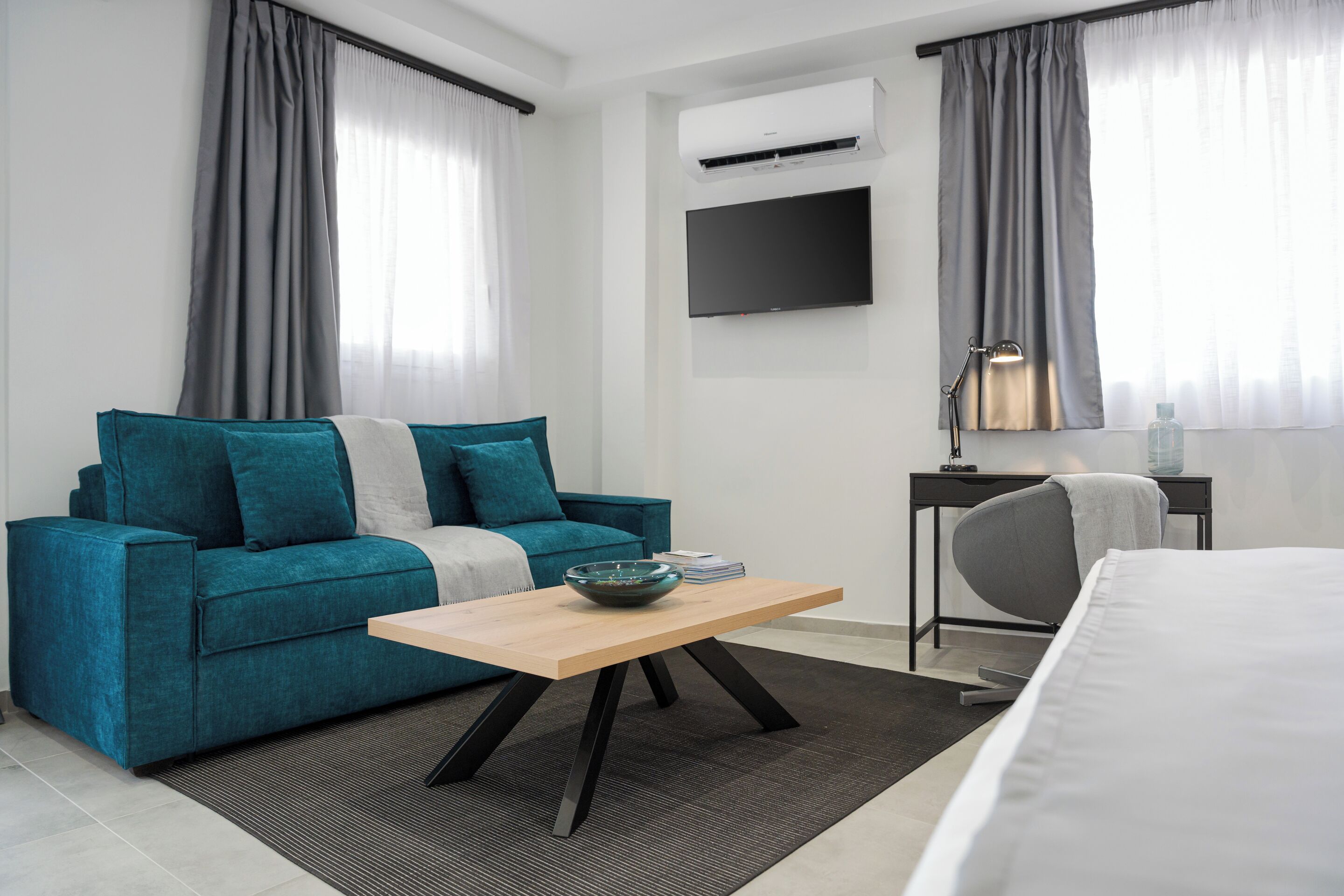 A modern hotel room with a teal sofa, a wooden coffee table, a bed, a desk with a lamp, and a TV mounted on the wall.