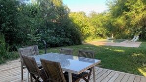 A wooden deck with a glass-top table and chairs, surrounded by lush greenery and a pool in the background.