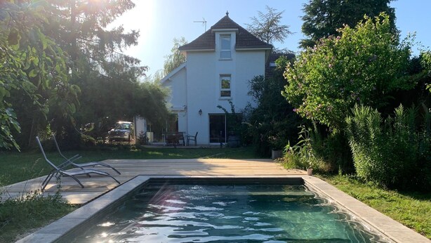 A swimming pool with a wooden deck and lounge chairs, surrounded by greenery and a house in the background.