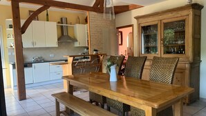A dining area with a wooden table and benches, a kitchen in the background, and a cabinet with glassware.