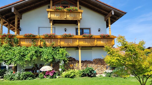 Un edificio de dos pisos con un balcón, un porche cubierto y un jardín con diversas plantas y flores.