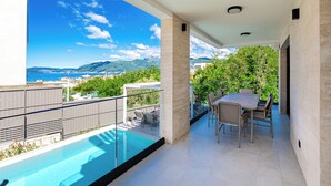 A balcony with a pool, outdoor seating, and a view of the sea and mountains.
