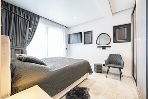 A modern bedroom with a large bed, a chair, a round mirror, and a window with curtains.