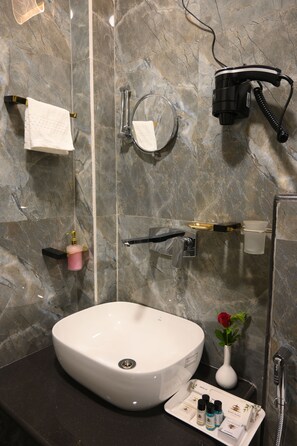 A bathroom with a white sink, a handheld hair dryer, a towel rack, a mirror, a rose in a vase, and toiletries on a tray.