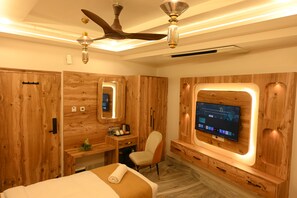 A hotel room with wooden paneling, a bed, a desk, a chair, a television mounted on the wall, and a ceiling fan. - Hotel Kashi Crown (Varanasi)