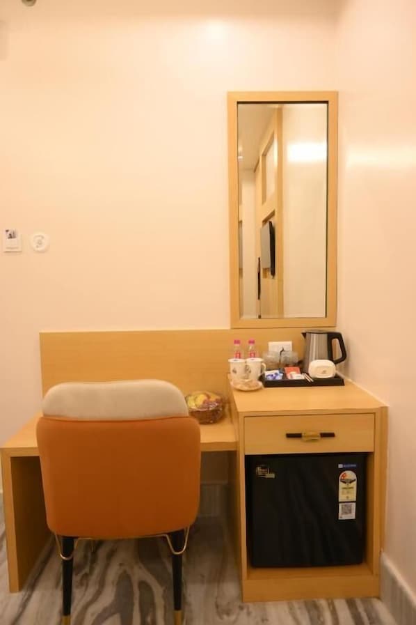 A compact hotel room with a wooden desk, a single orange chair, a mirror, and a small cabinet with a black interior.