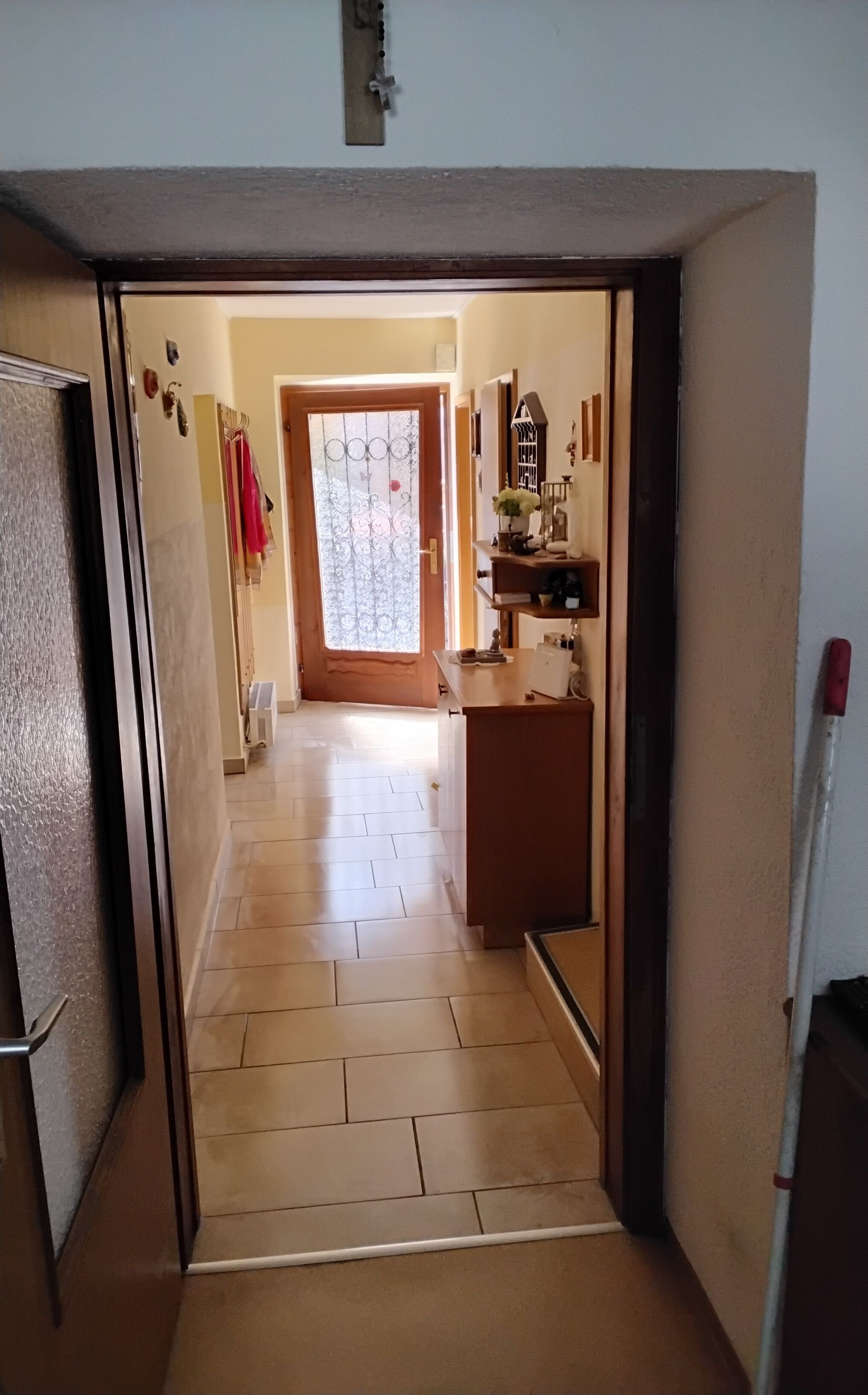 A narrow hallway with a wooden door on the left, a small kitchen area with a sink and counter on the right, and a view into a room with a window and a door at the end.