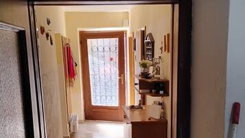 A narrow hallway with a wooden door on the left, a small kitchen area with a sink and counter on the right, and a view into a room with a window and a door at the end.