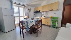 Een kleine keuken met een eettafel, stoelen, een koelkast, houten kastjes, een gootsteen en een groene opbergwagen.