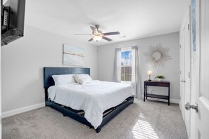 Ein Schlafzimmer mit Bett, Deckenventilator, Fenster mit Vorhängen, Nachttisch mit Lampe und einem Wandspiegel.