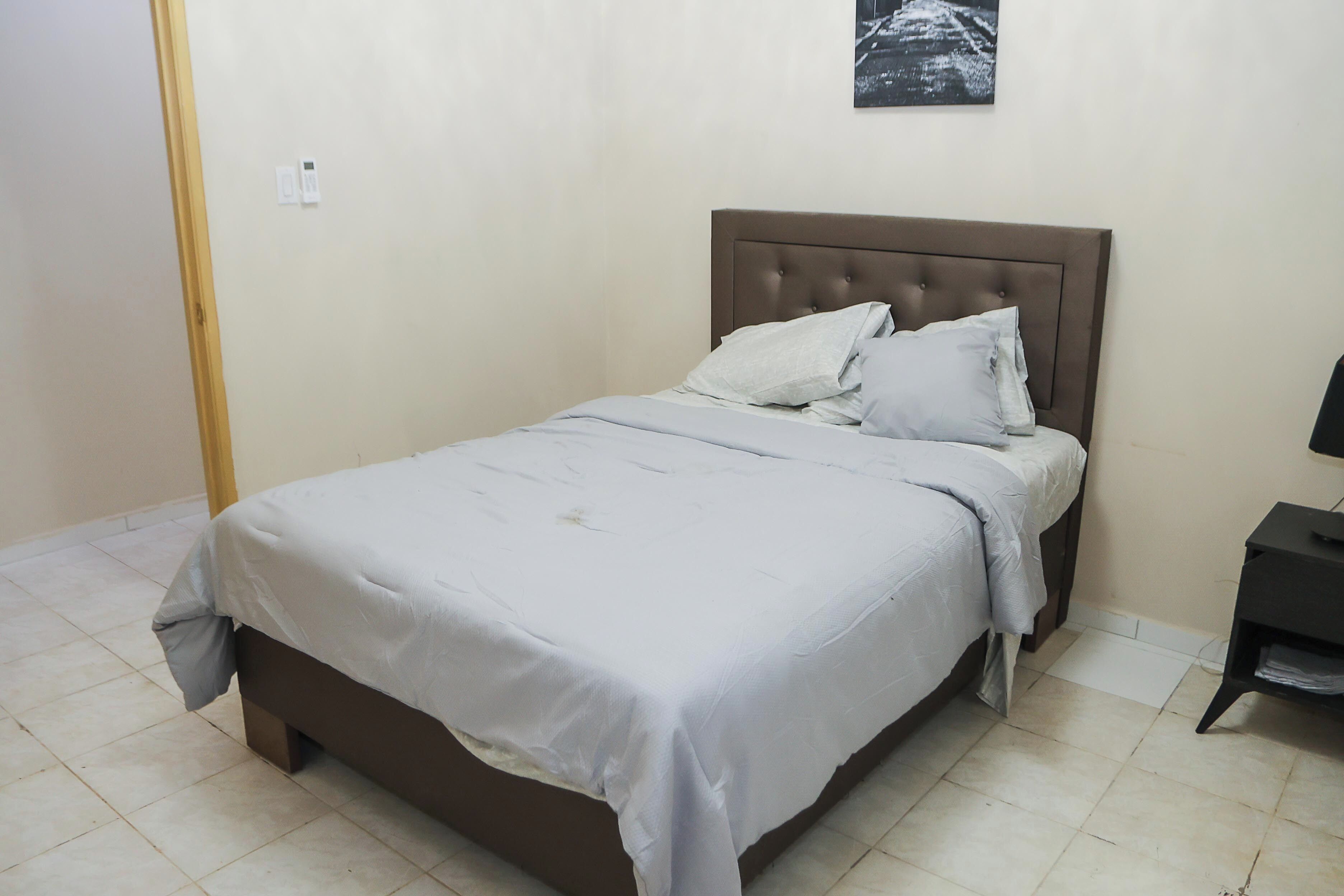 Cama bem arrumada com colcha cinzenta e almofadas, cabeceira de madeira e uma pequena mesa de cabeceira com abajur.