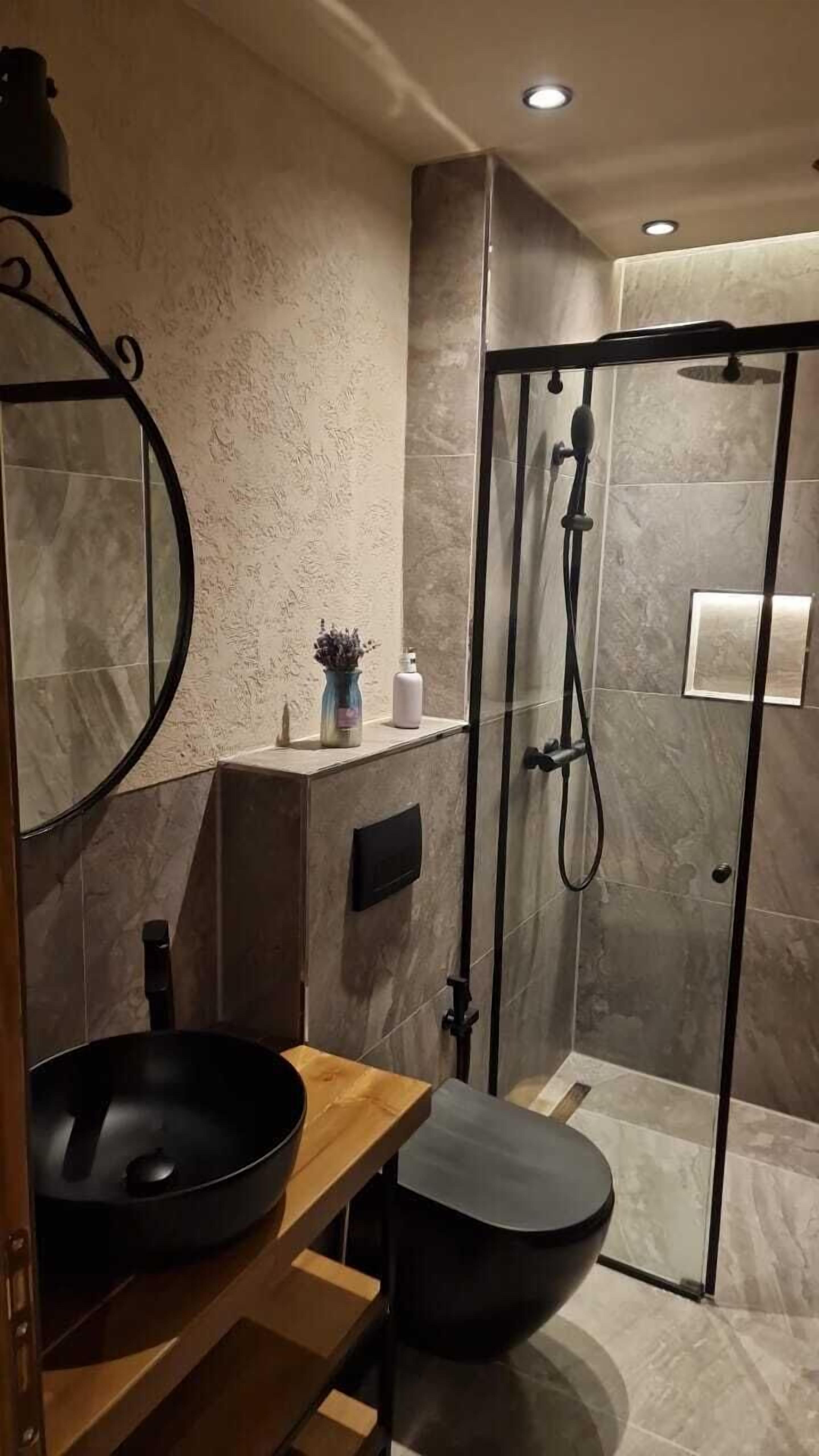 Un baño moderno con un espejo redondo, un lavamanos negro, un inodoro y una ducha con mampara de vidrio.