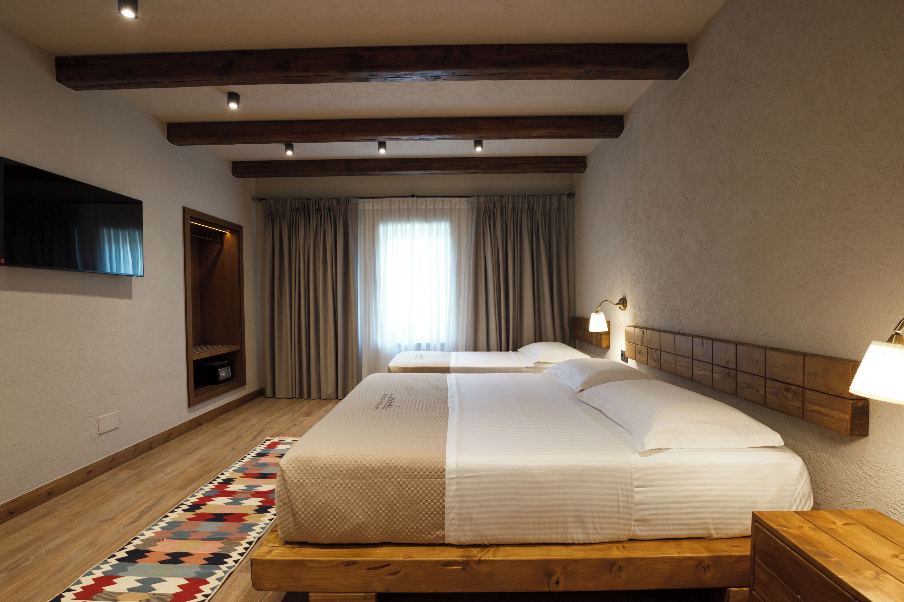 Een hotelkamer met twee bedden, een aan de muur bevestigde televisie, een houten hoofdbord, een nachtkastje met een lamp en een kamerkleed met dessin.