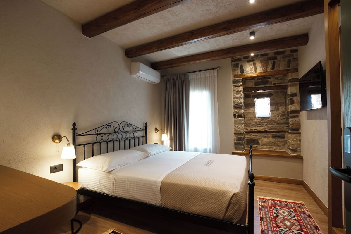 A bedroom with a bed, a nightstand, a lamp, a stone wall, and a wooden ceiling beam.