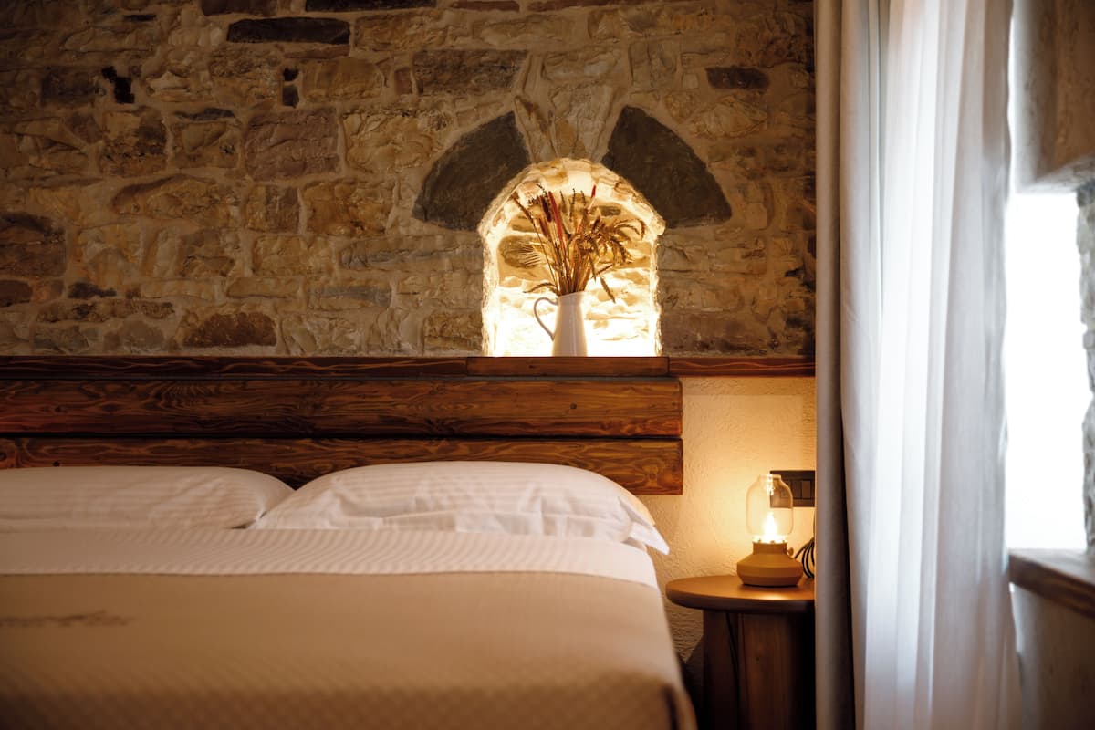 A room with a stone wall, a wooden headboard, a bed with white bedding, and a small table with a lamp.