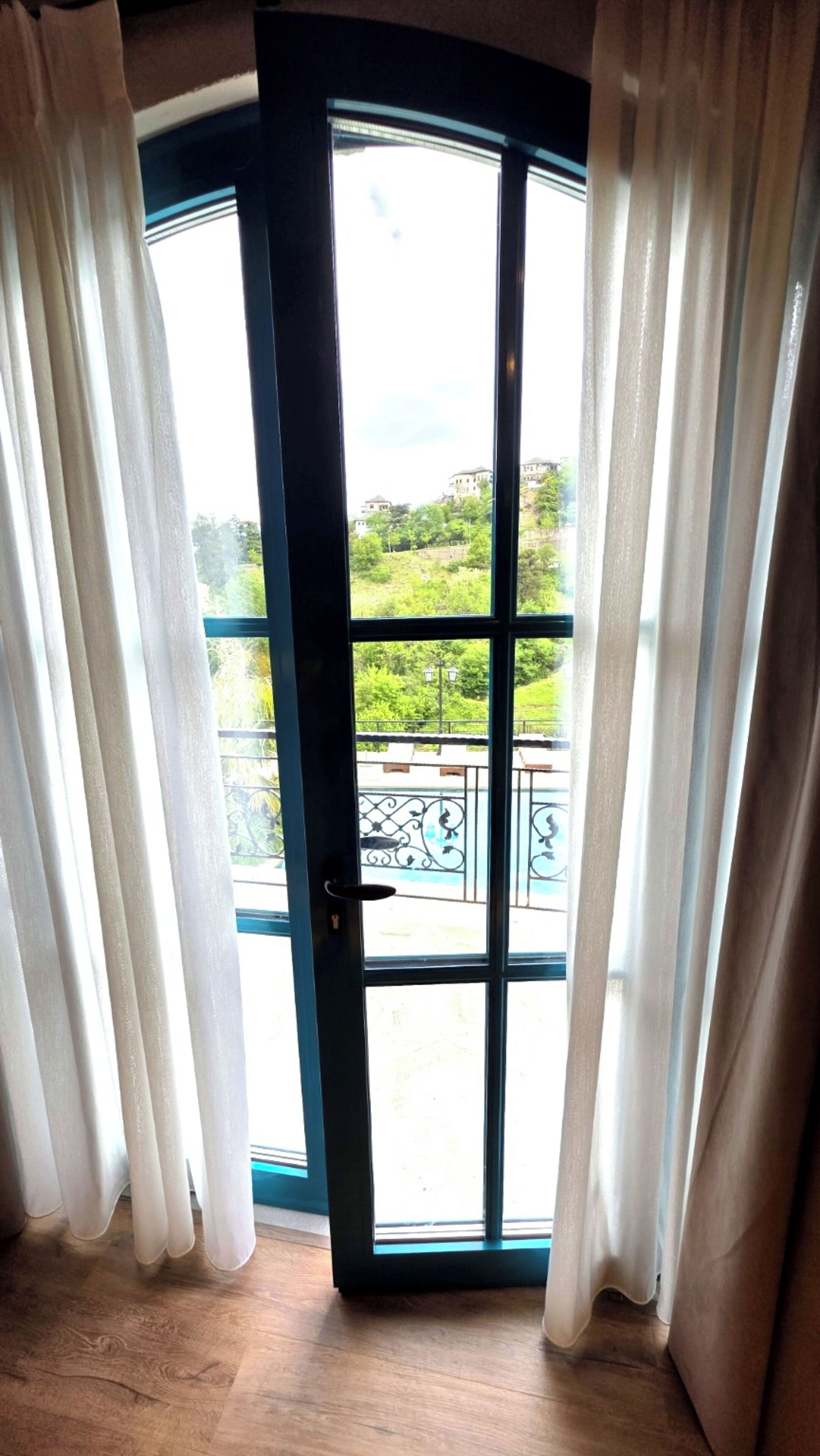 A room with a large glass door leading to a balcony with a metal railing and greenery in the background.
