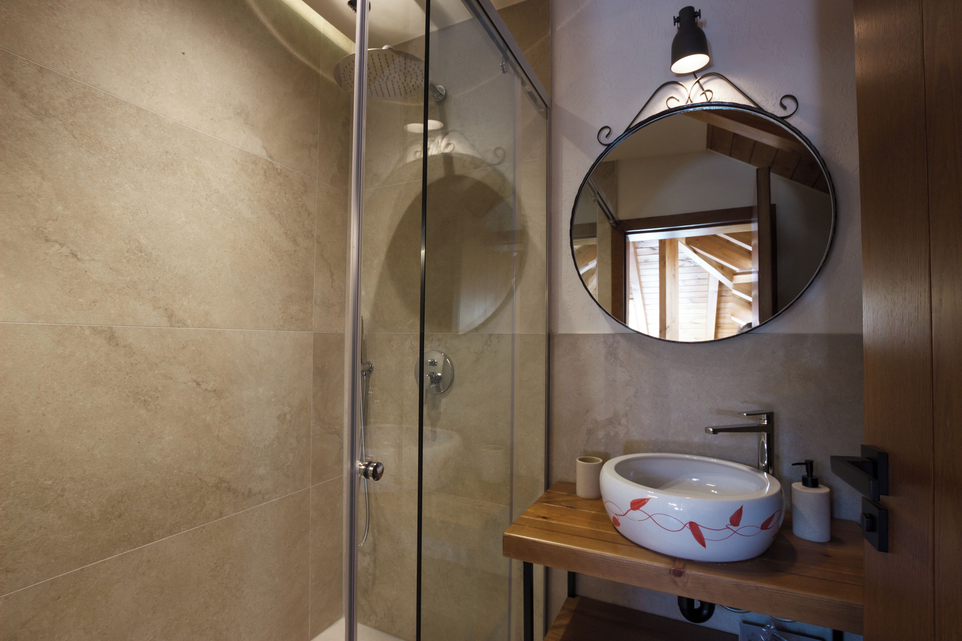 A bathroom with a glass-enclosed shower, a wooden vanity, a round mirror, and a white sink with a red leaf design.