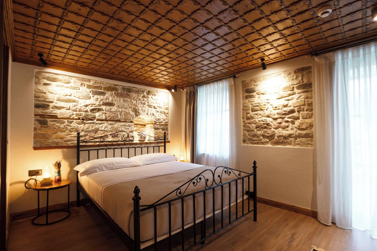 A bedroom with a stone wall, a bed with a metal frame, a small bedside table, and a window with curtains.