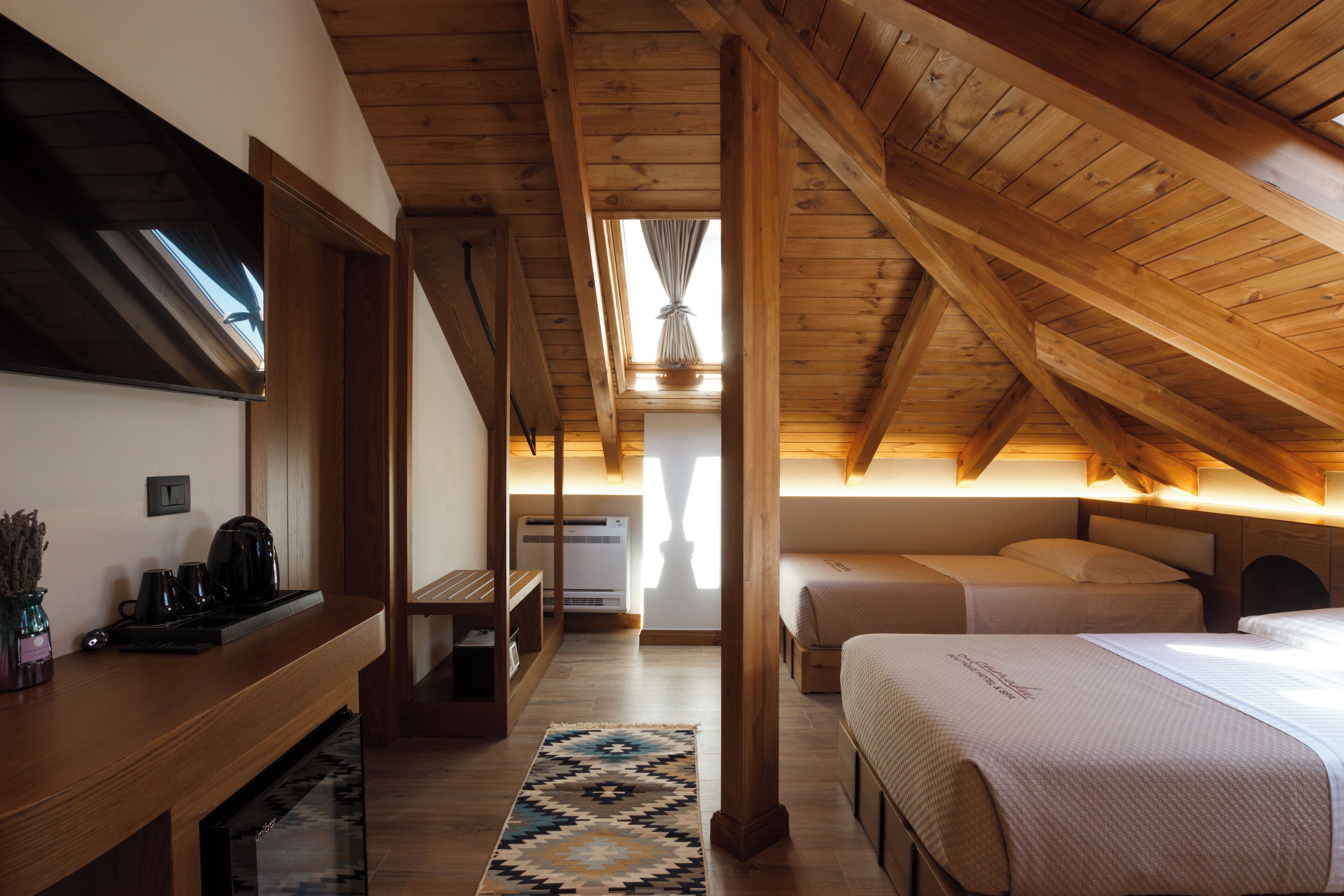Une chambre avec un plafond en pente, des poutres en bois, un lit, des tables de chevet, un bureau et une fenêtre avec des rideaux.