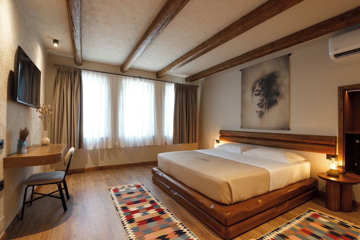 A bedroom with a large bed, wooden beams, a mounted TV, a painting on the wall, and a window with curtains.